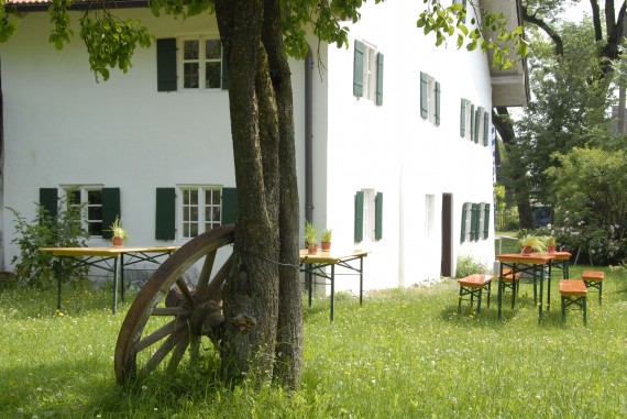 Haus und Garten des Weiß-Egger-Anwesens in Ellighofen, ein großes Wagenrad lehnt am Baum, Biertische stehen im Garten.