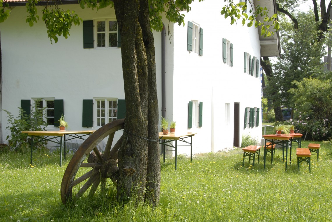 Weißes Haus und Garten des Weiß-Egger-Anwesens in Ellighofen, ein Wagenrad lehnt am Baum, Bierbänke stehen auf der Wiese.
