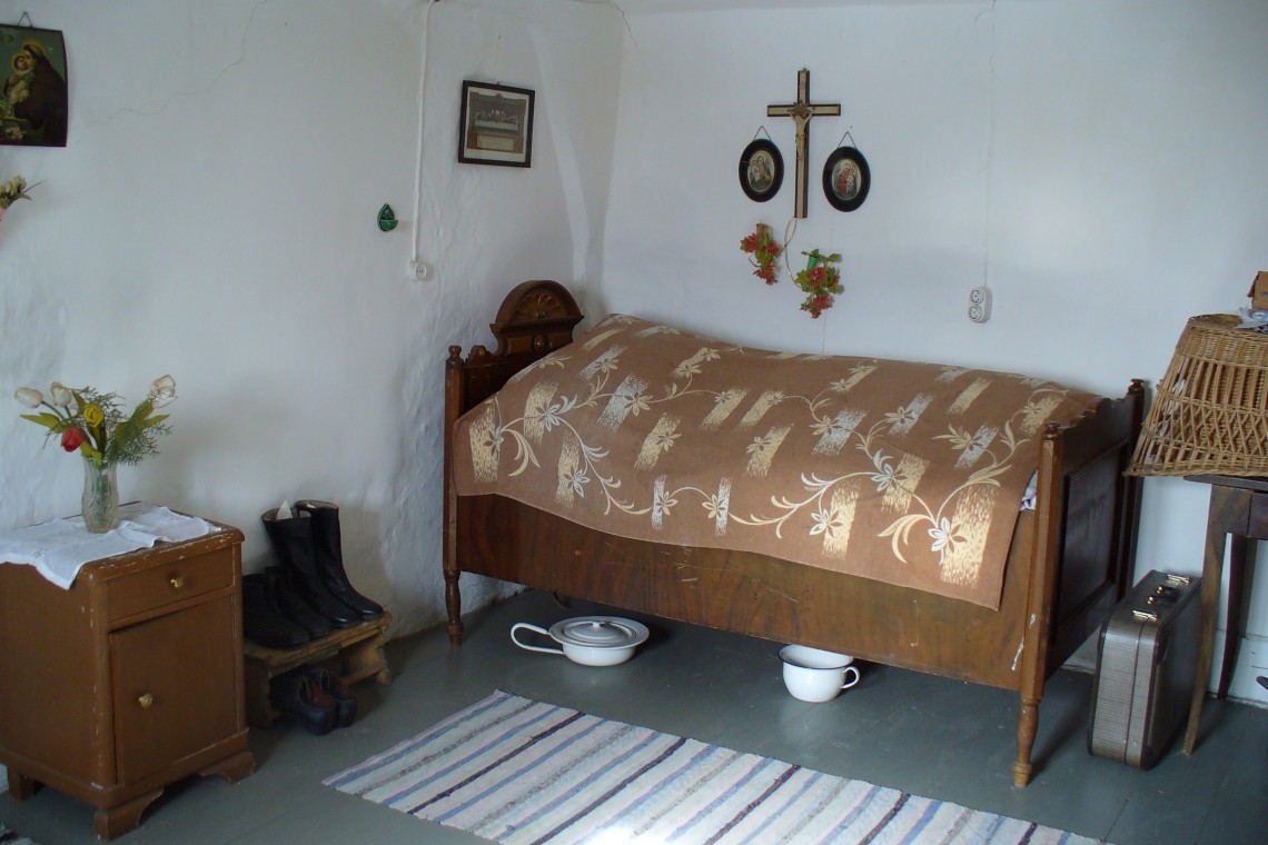 Schlafzimmer im Bauernhaus mit einfachster Einrichtung: ein schmales Bett, darüber ein Kruzifix und Heiligenbilder, unter dem Bett eine Bettpfanne. 