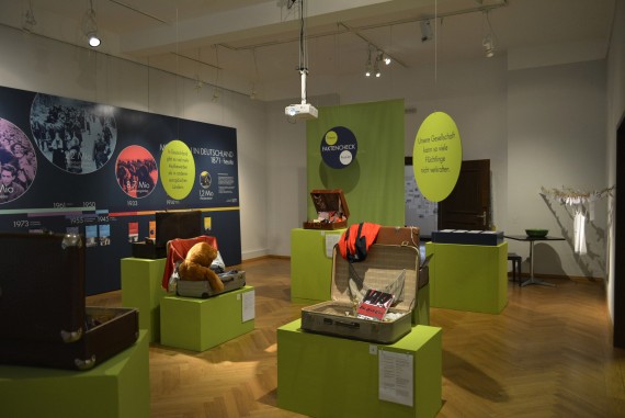 Blick in die Migrations-Ausstellung, Aufgeklappte Koffer auf hellgrünen Kästen, im Hintergrund eine Übersichtswand