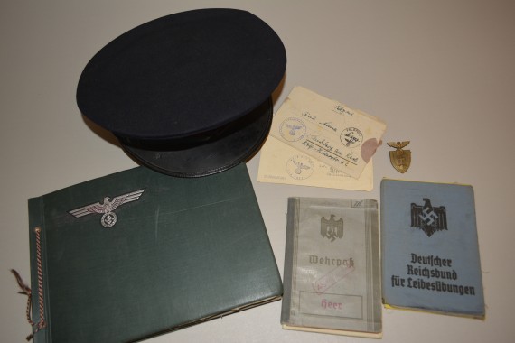Objekte auf einem Tisch angeordnet, eine Mütze der Deutschen Arbeiterfront, ein Fotoalbum, ein Wehrpass, ein Heft des Deutschen Reichsbund für Leibesübungen, ein goldfarbener Anstecker