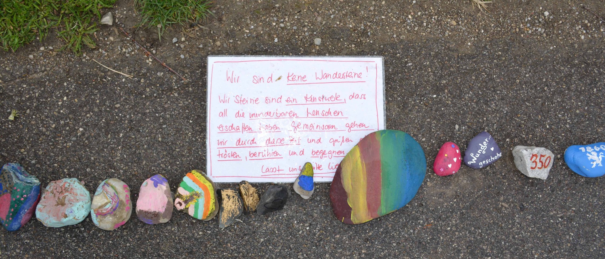 Eine Reihe bunt bemalter Steine am Straßenrand mit handgeschriebenem Text zur Idee.