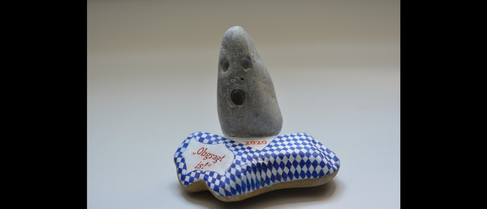 Skulptur aus zwei Steinen. Auf einem flachen Stein mit blau-weißer Rautenbemalung und der Aufschrift &quot;Obgsagt is!&quot; steht ein länglicher Stein mit drei Vertiefungen, die aussehen, wie ein schreiendes Gesicht.
