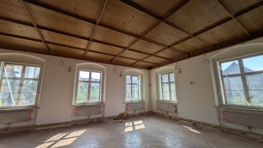 Raumecke mit großen Fenstern, durch die Licht hereinfällt. An der Decke sind Holzkassettenfelder zu sehen.