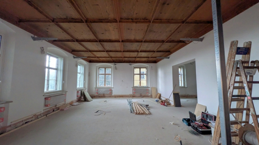 Großer heller Raum, an der Decke eine hölzerne Kassettendecke, an den Wänden oben graue Metallausleger.