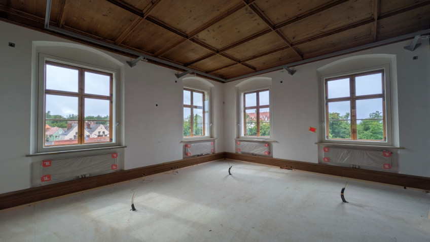 Raum mit großen Fenstern und Holzkasettendecke, aus dem Boden ragen Kabelführungen.