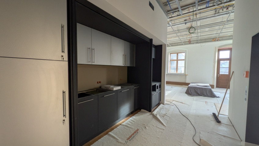 Links im Raum ist eine dunkelgraue K&uuml;chenzeile in eine Nische eingebaut.